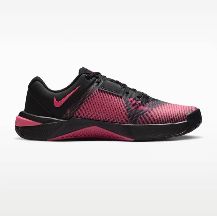 Профилен изглед на дамска обувка за тренировки Nike Metcon 10 SE. Обувката е с черна основа, дишаща мрежа с розов преливащ десен в предната част, розово лого Nike Swoosh на петата и здрава подметка с розови детайли.